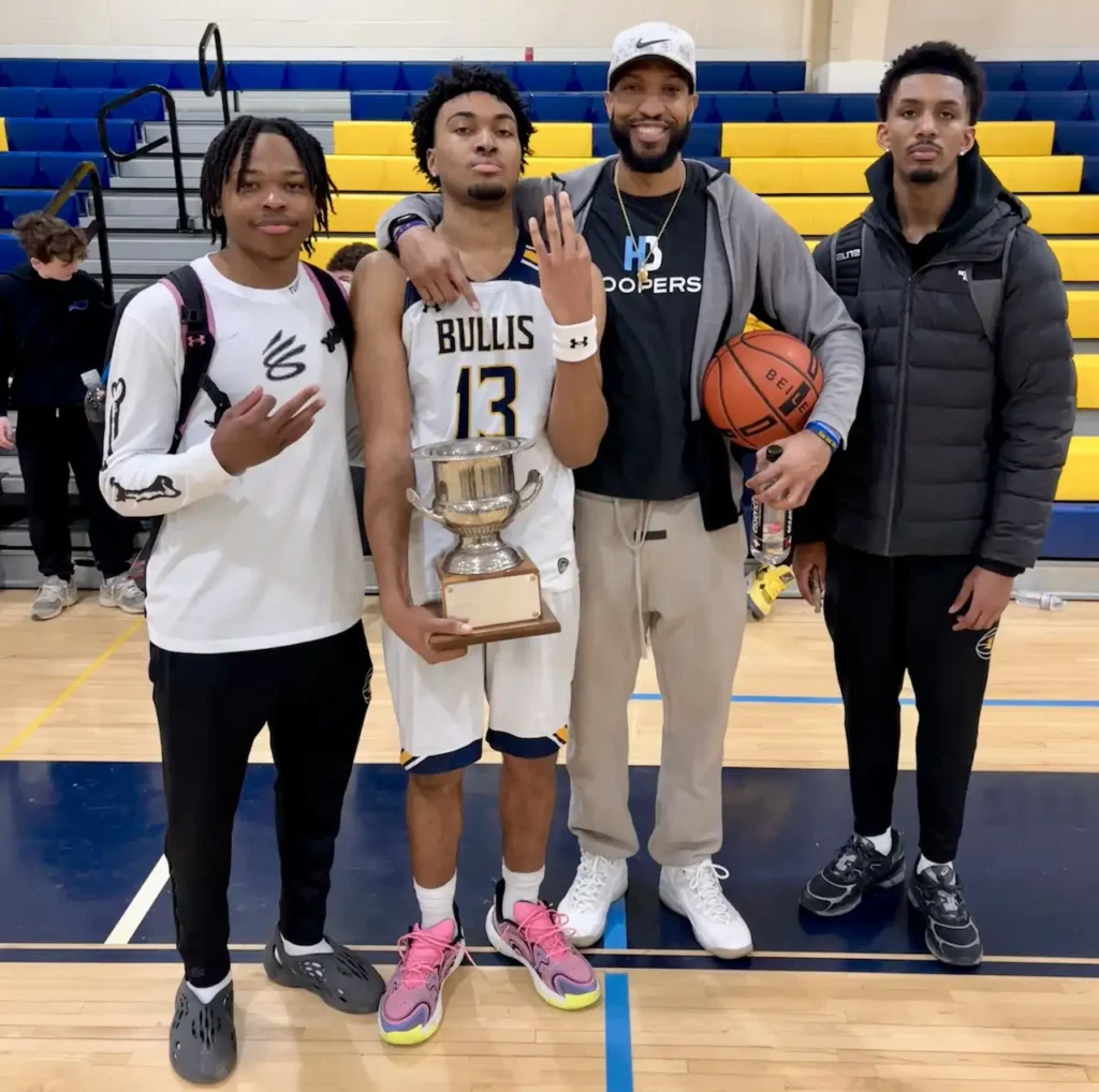 Four basketball players celebrate with trophy on court.
