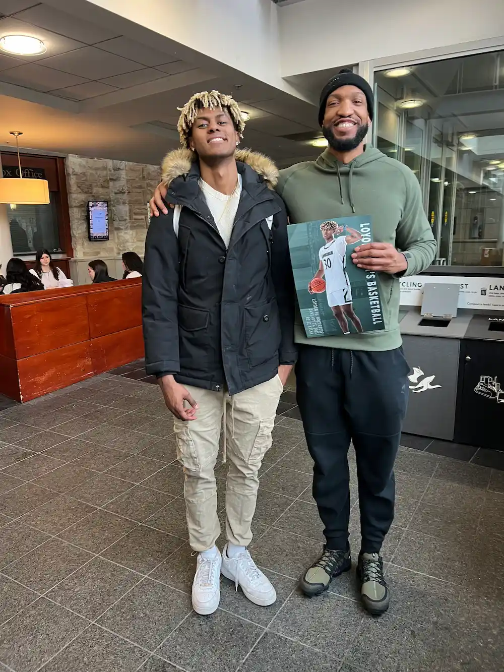 Two men smiling in a hallway with basketball poster.